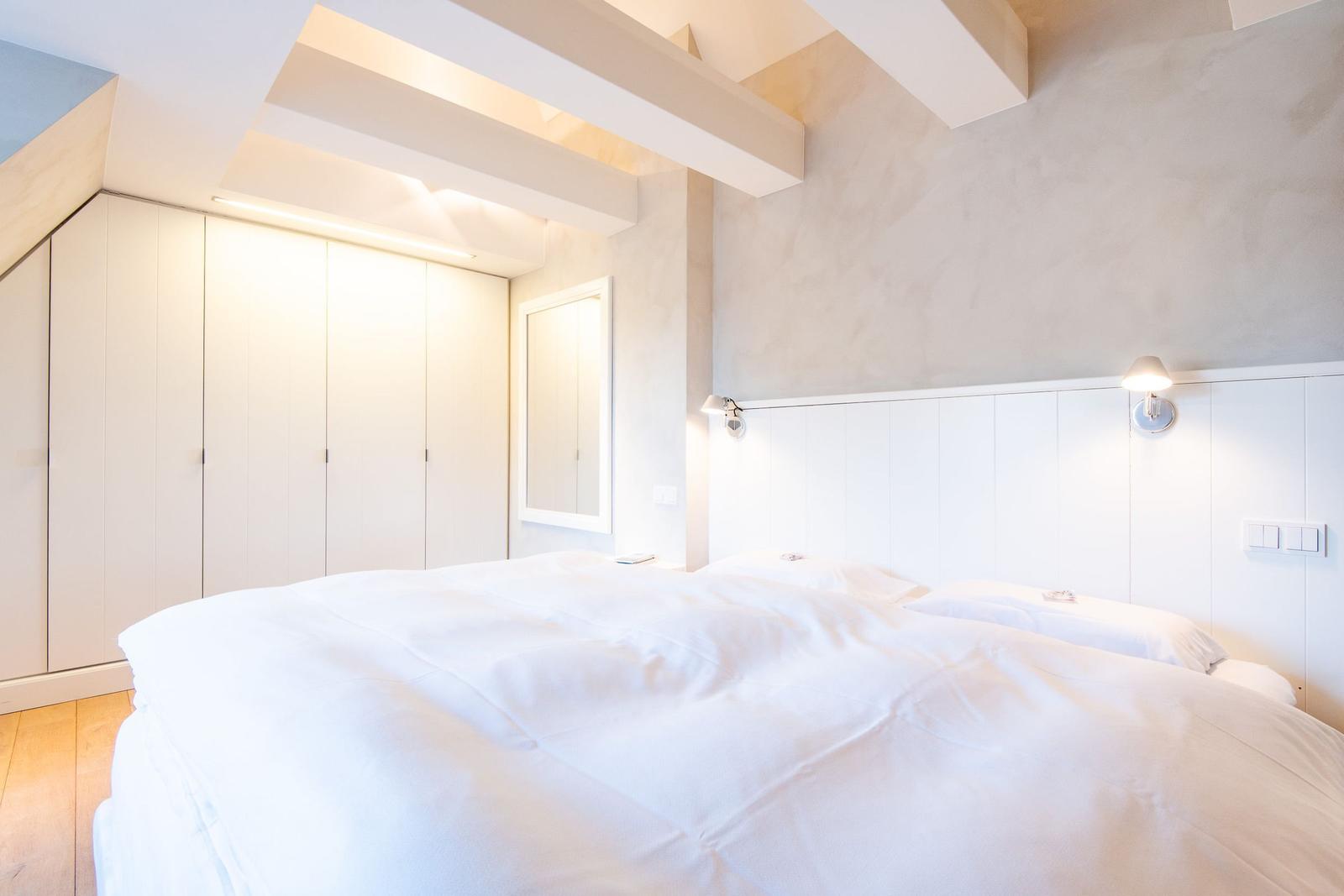 Schlafzimmer mit weißem Bett, Kleiderschrank und Wandlampen unter Holzbalkendecke.