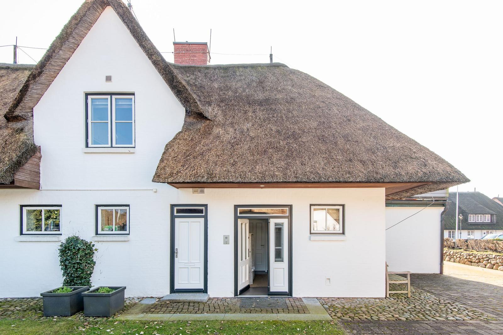 Wohnhaus mit Strohdach, weißen Wänden und zwei Eingängen. Vorgarten mit Pflanzen.