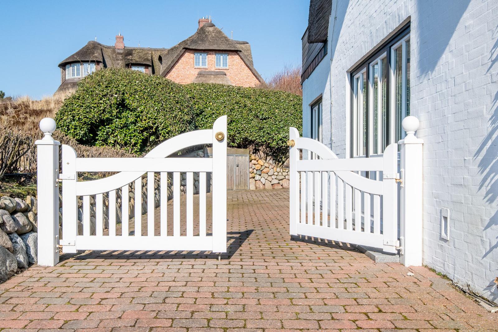 Weiße Torflügel öffnen den Blick auf einen gepflasterten Weg und ein traditionelles Haus mit Strohdach.