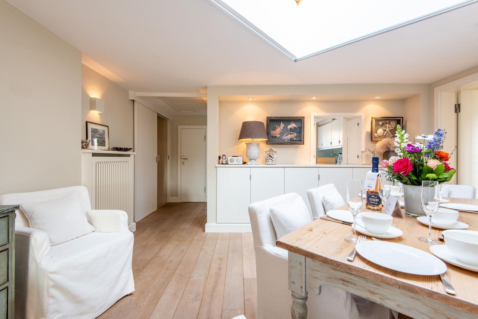 Essbereich mit Holztisch und weißen Stühlen. Hintergrund zeigt Küchenbereich und Wohnbereich.