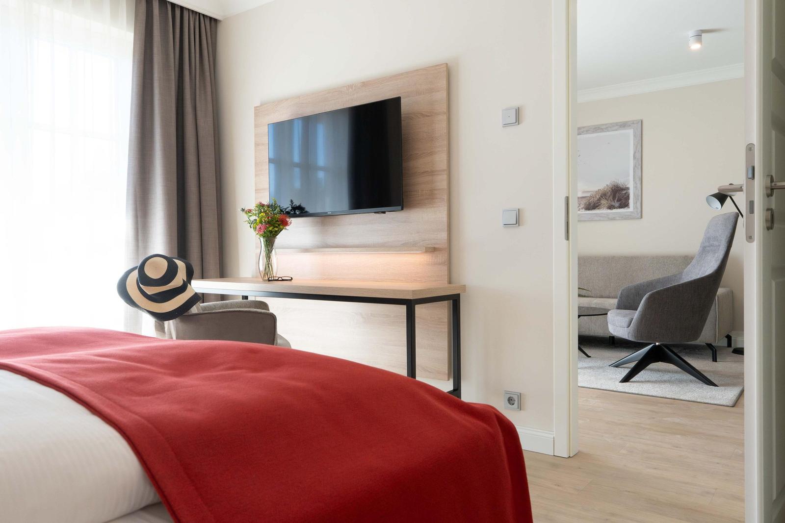 Schlafzimmer mit rotem Bettbezug, Fernseher und Sicht auf Wohnzimmer.