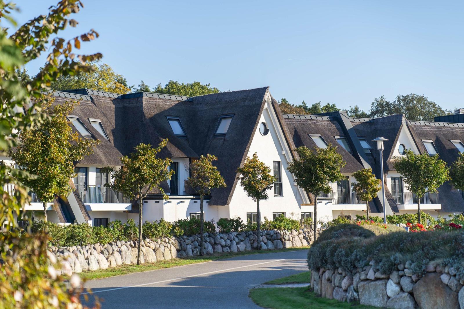 Reihenhäuser mit Strohdächern und Balkonen entlang einer Straße mit Steinmauern und Bäumen.