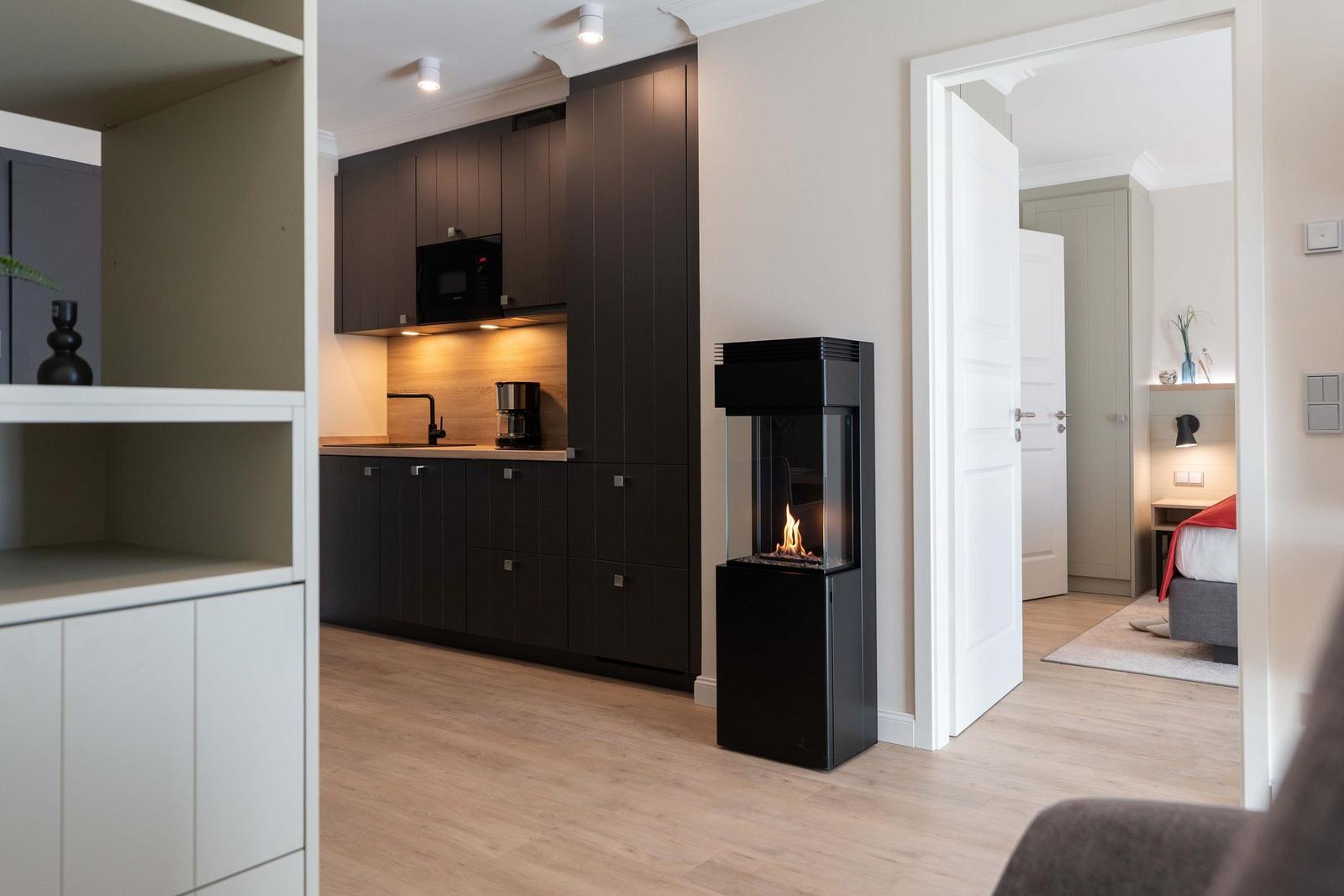 Küche mit schwarzen Schränken und Kamin im Wohnbereich. Schlafzimmer ist durch Tür sichtbar.