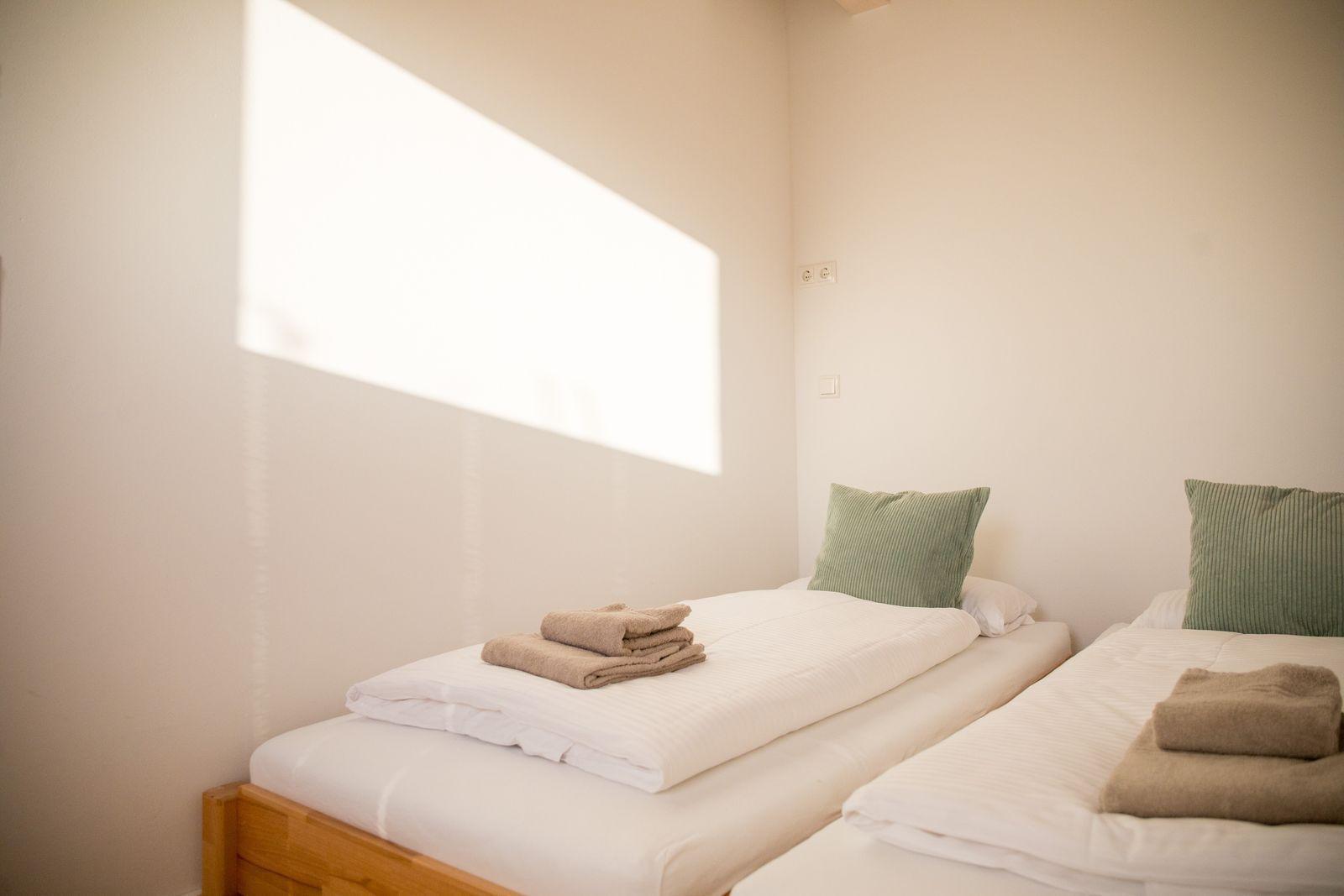 Zwei Betten mit weißen Bettwäsche und grünen Kissen. Ein Fenster lässt Licht herein.