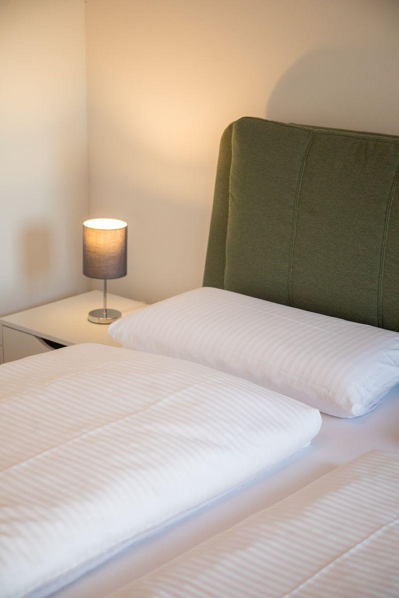 Ein Bett mit weißer Bettwäsche und grünem Kopfteil. Neben dem Bett steht ein Nachttisch mit einer Lampe.