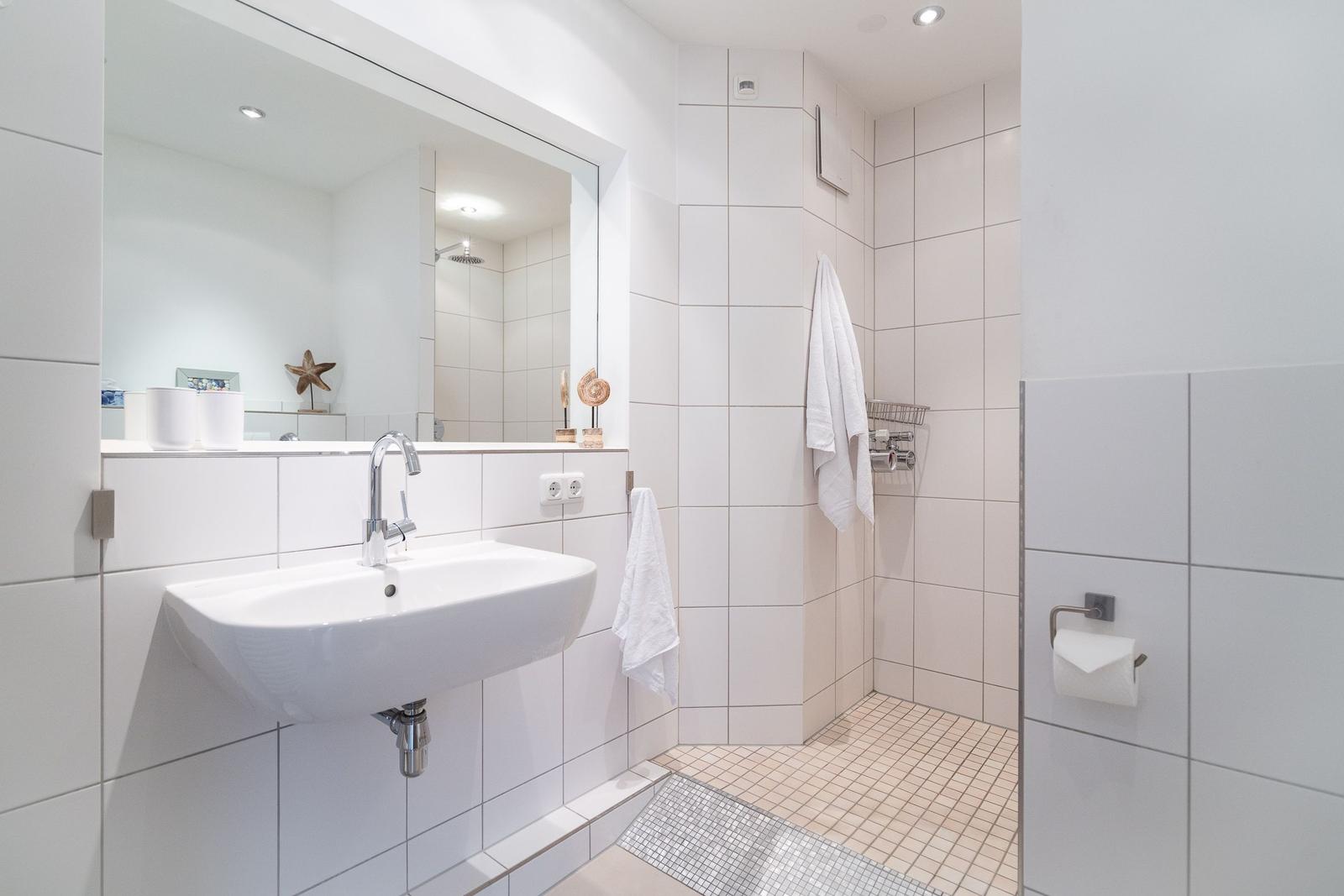 Badezimmer mit Waschbecken, Spiegel und Dusche. Weiße Fliesen und Handtuchhalter.