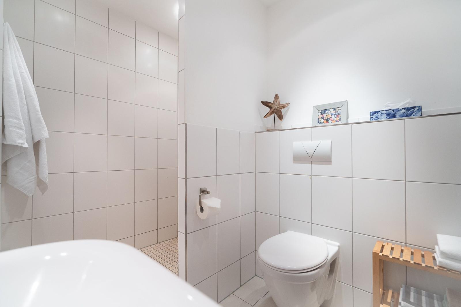 Badezimmer mit Toilette, Dusche und Badewanne. Weiße Fliesen und Holzregal.