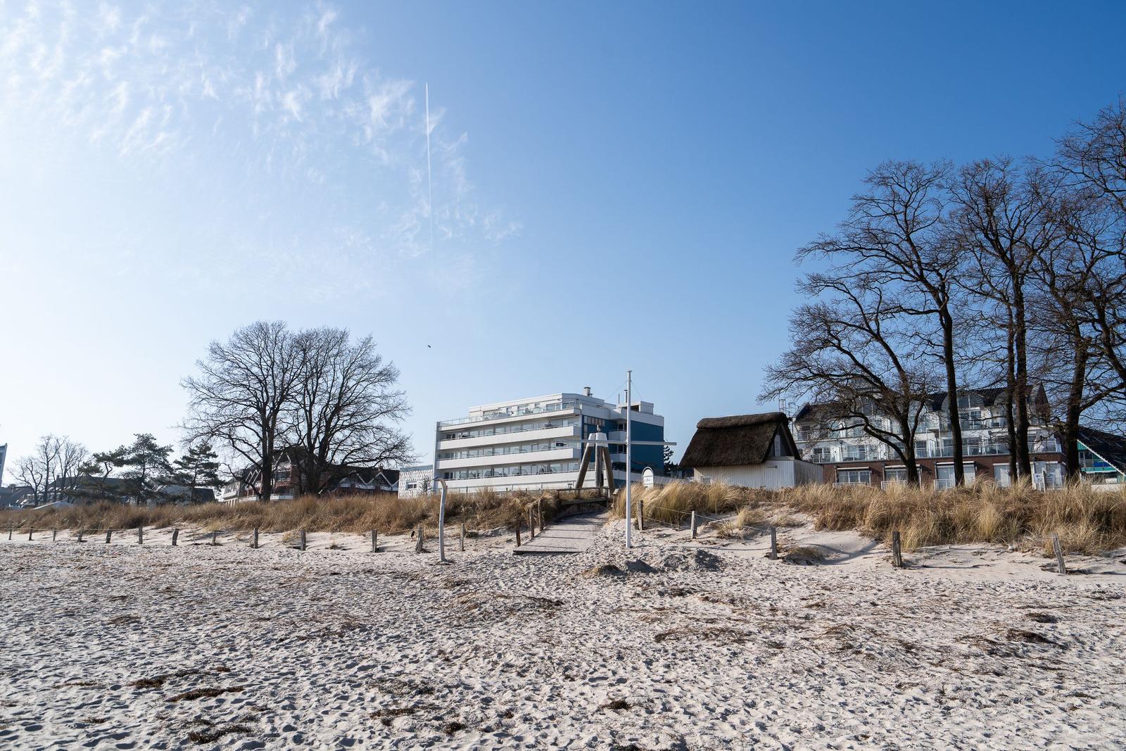 Strand mit Dünen, Bäumen und Gebäuden im Hintergrund.