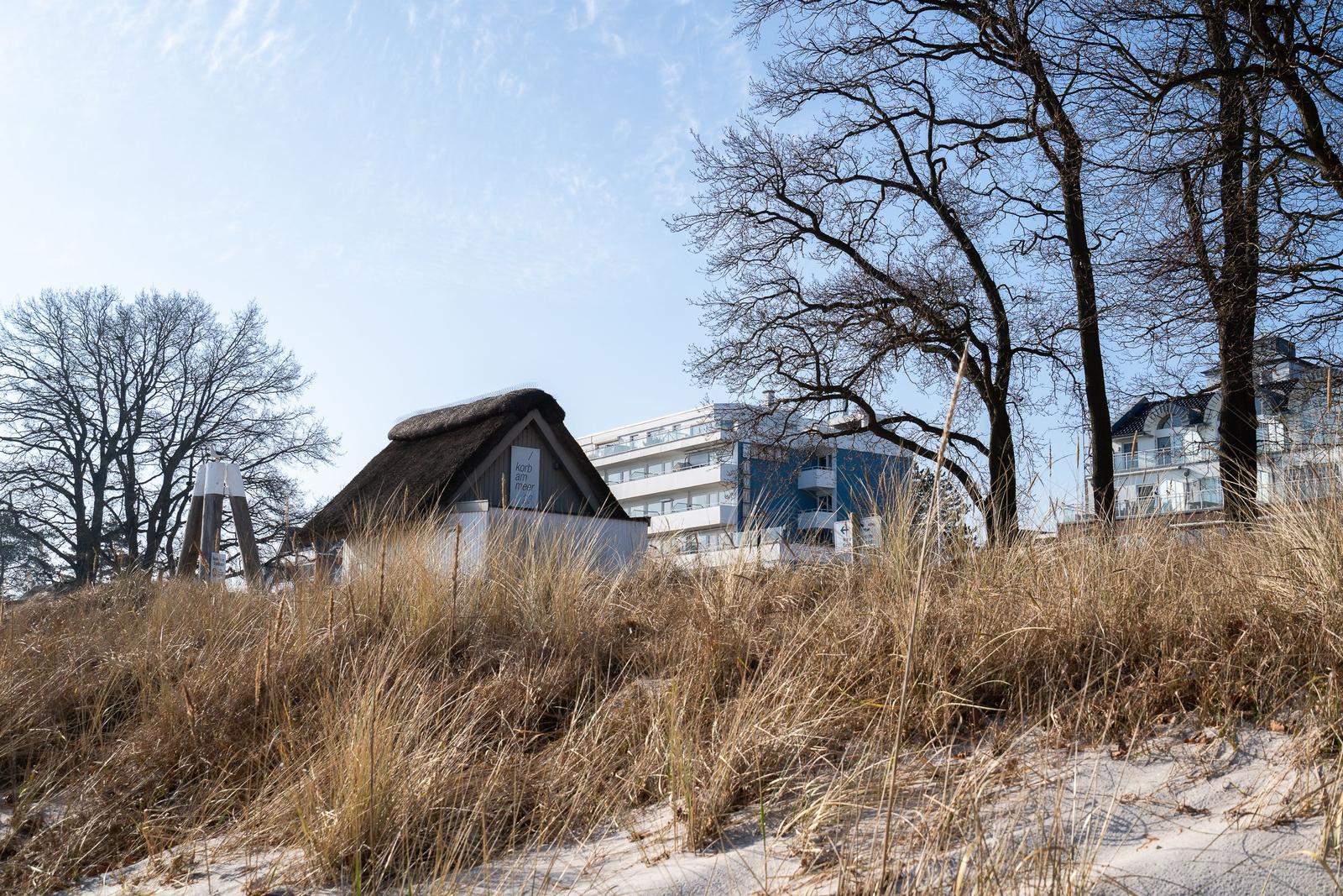 Dorfhaus mit Strohdach auf Dünen, moderne Gebäude im Hintergrund.