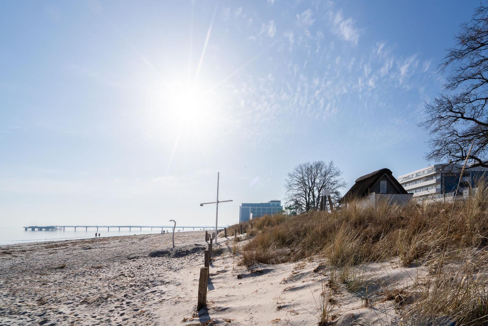 Strand mit Dünen, Hütte und Pier unter blauem Himmel.