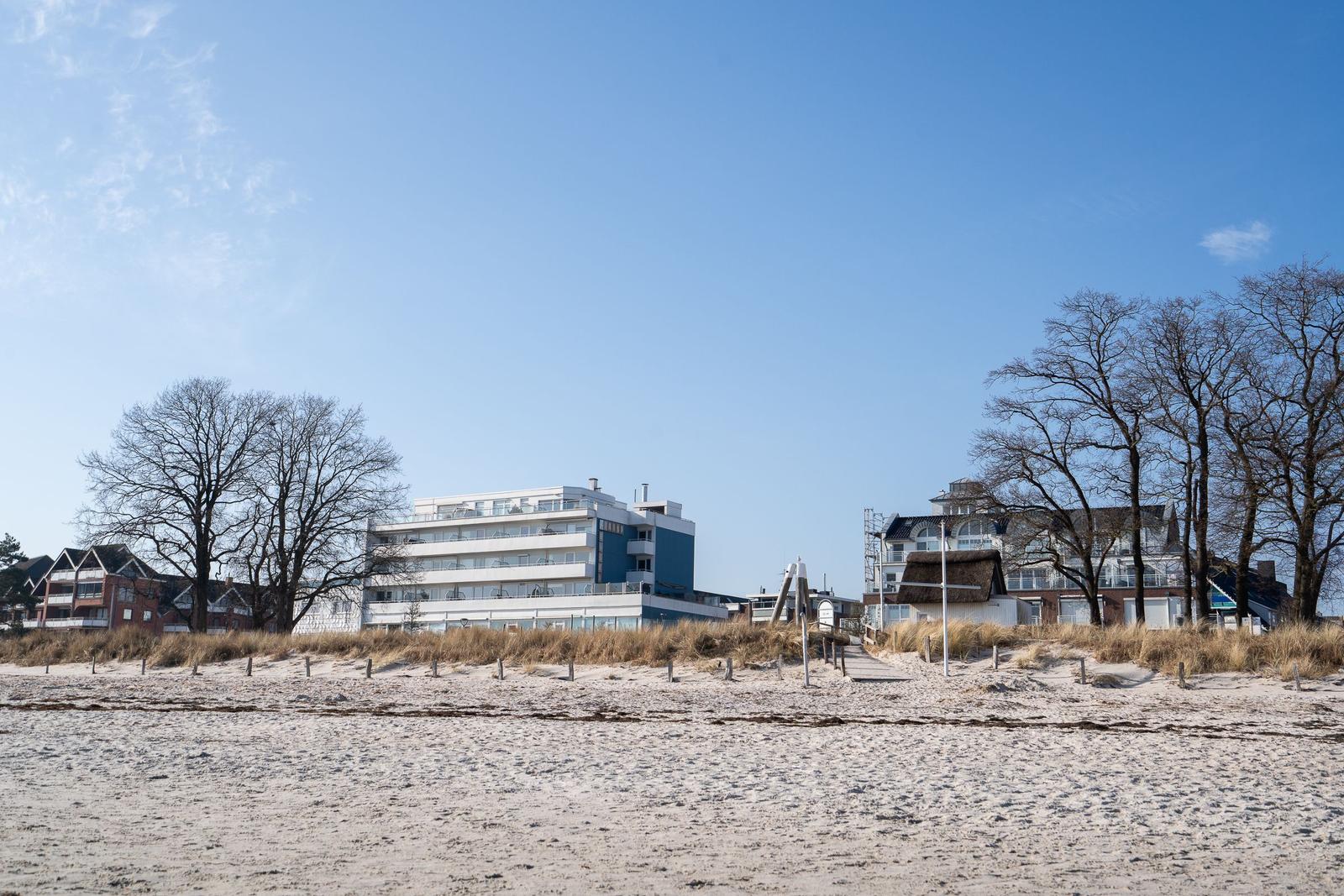 Strand mit Dünen, Bäumen und Ferienhäusern im Hintergrund unter blauem Himmel.