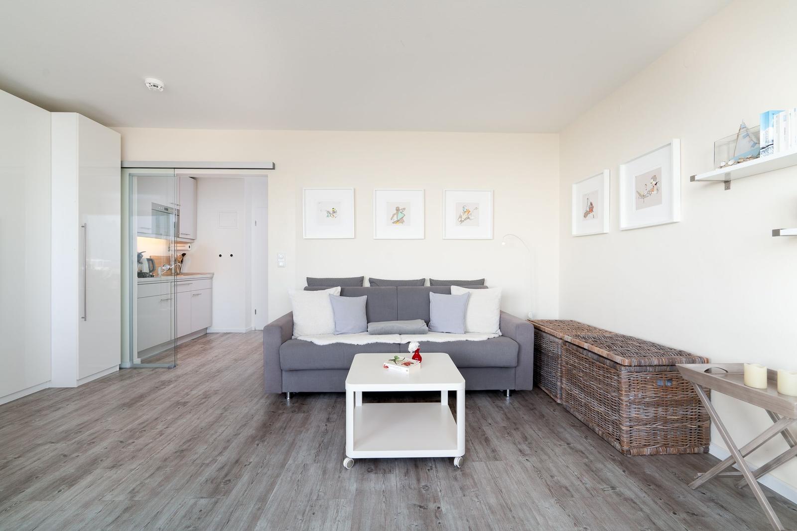 Wohnbereich mit grauem Sofa, weißen Kissen und Holzboden. Küche im Hintergrund.