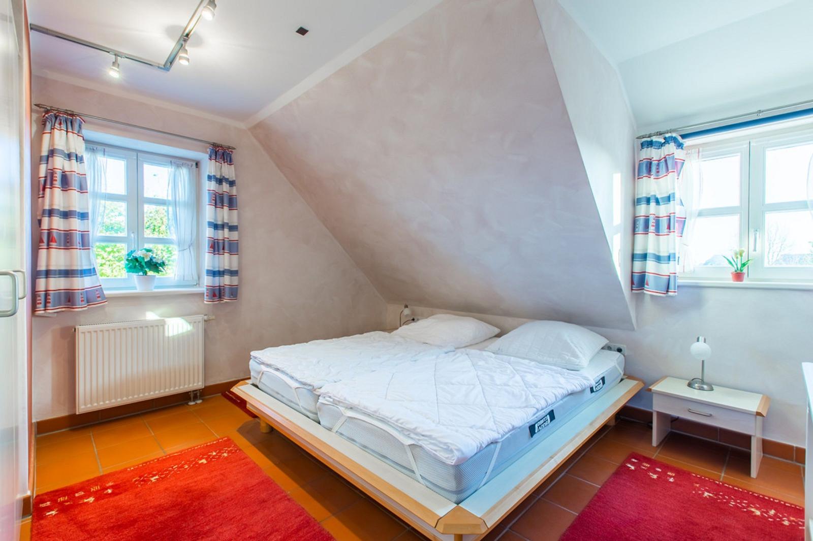 Schlafzimmer mit Dachschrägen, Bett, zwei Fenstern und Teppichen.