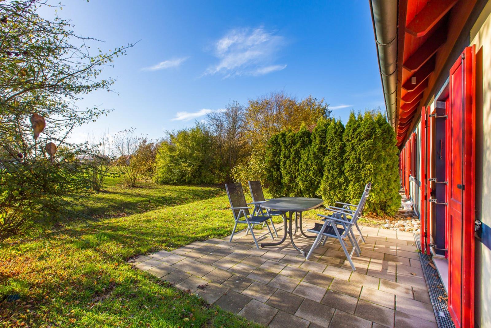 Terrasse mit Tisch und Stühlen neben roten Türen. Grünflächen und Bäume im Hintergrund.