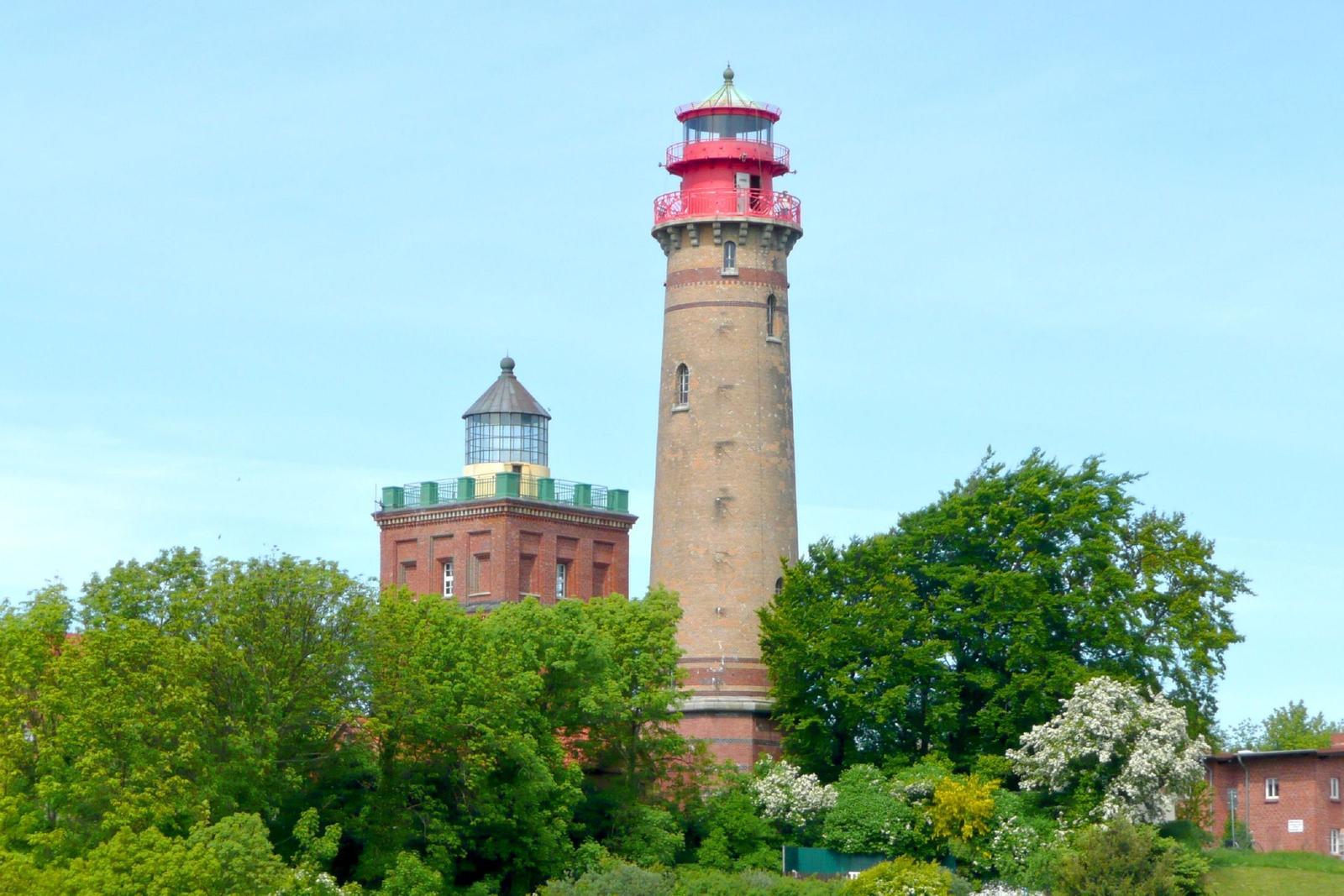 Zwei Leuchttürme stehen unter blauem Himmel, umgeben von grünen Bäumen.