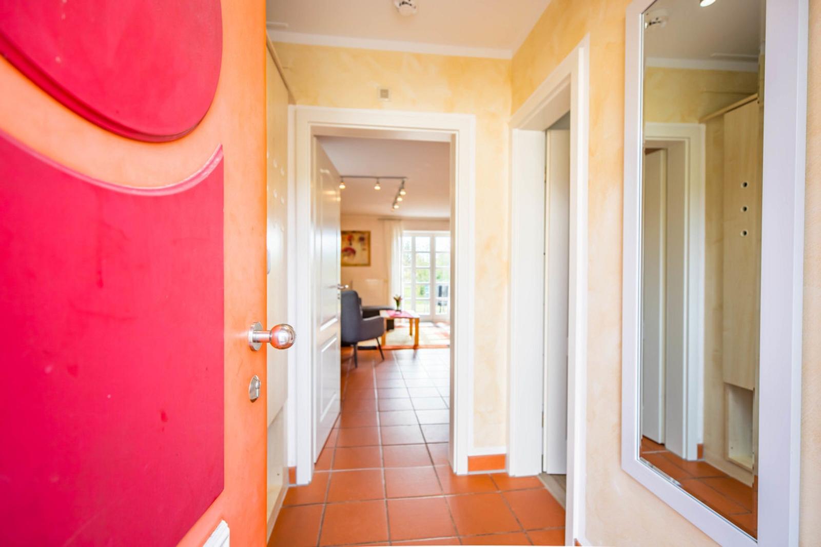 Hausflur mit orangefarbener Tür und Spiegel. Blick in Wohnzimmer mit Sitzgelegenheit.