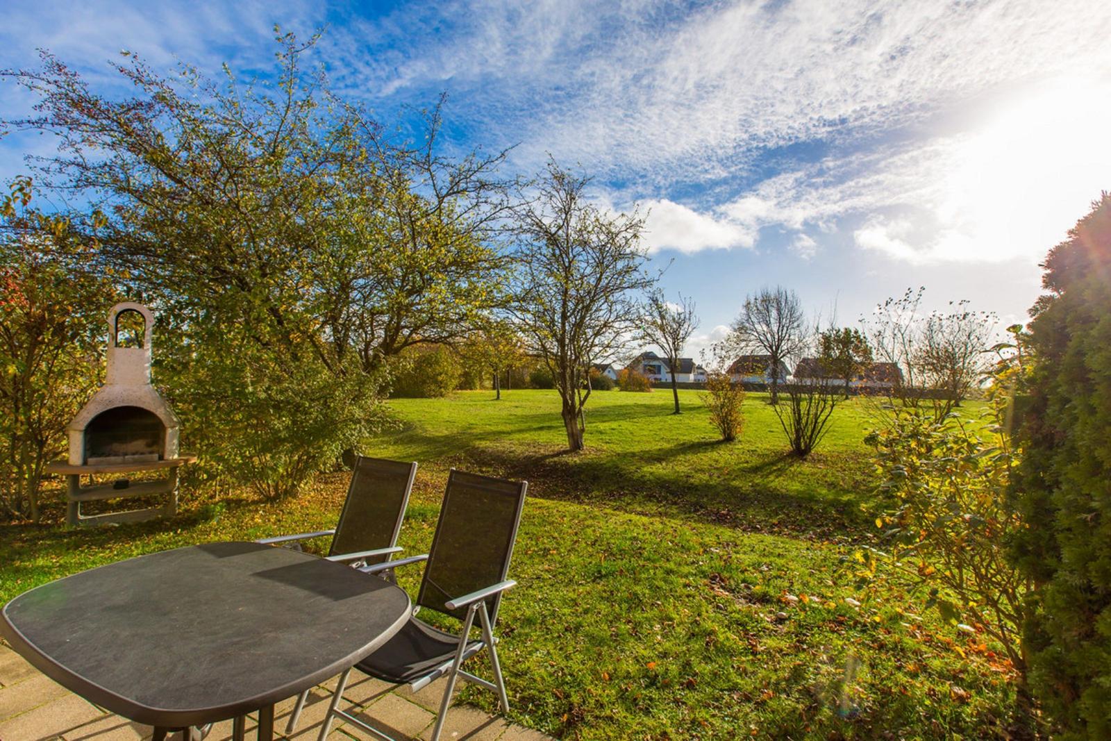 Außenbereich mit Tisch, zwei Stühlen und Grill. Grünfläche mit Bäumen und Häusern im Hintergrund.