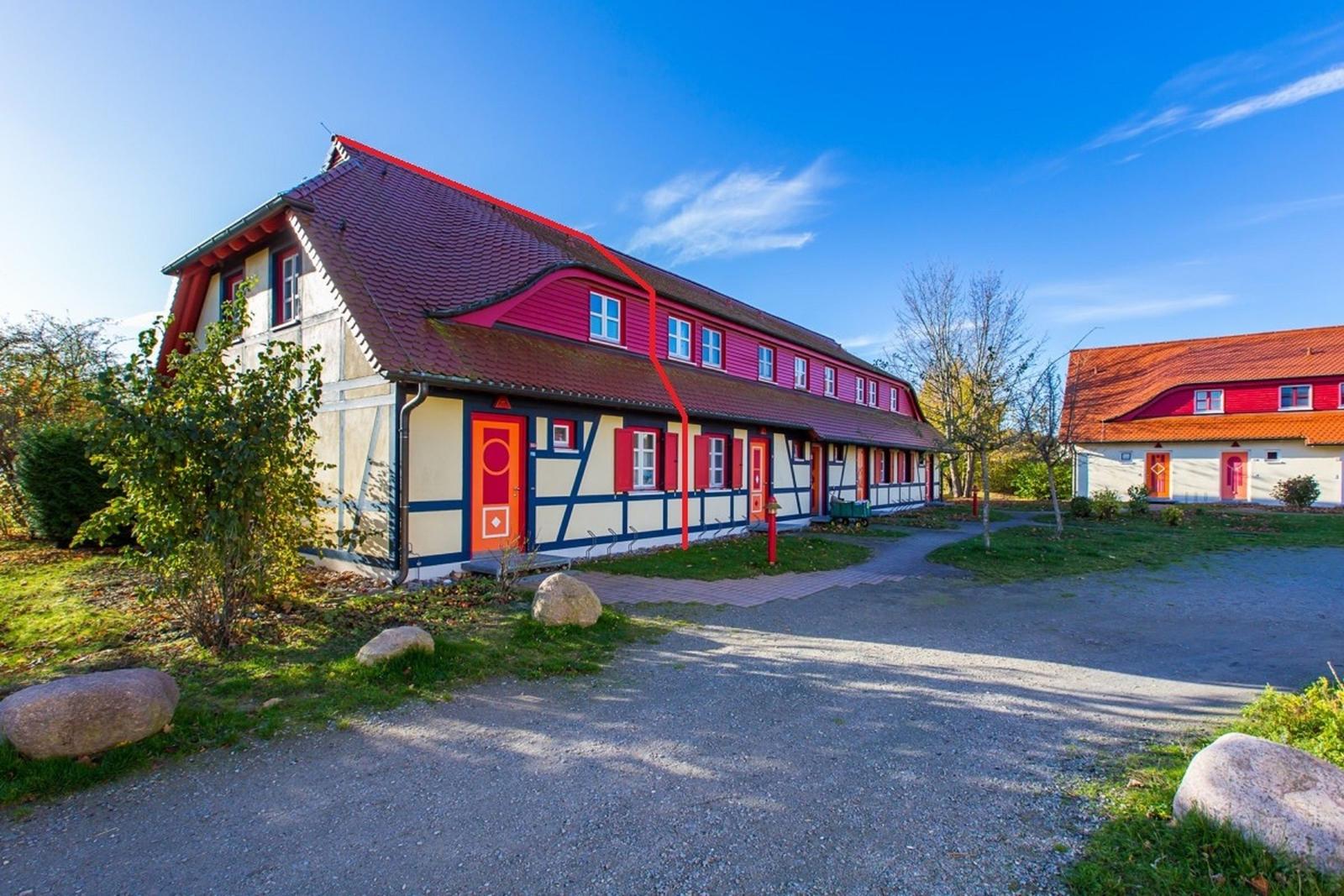 Ferienhaus mit Fachwerk, rotem Dach und orangefarbenen Türen. Grünflächen und Steine im Vorgarten.