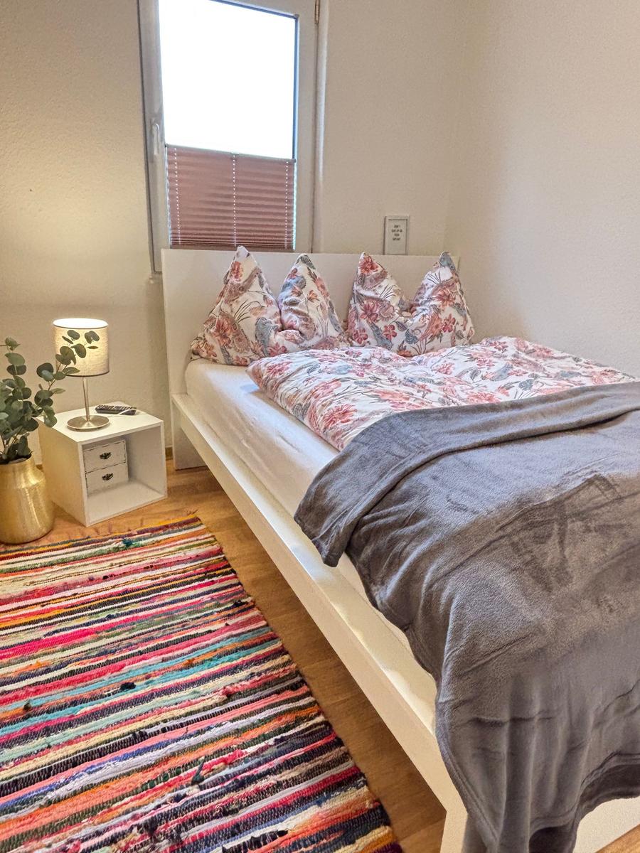 Schlafzimmer mit Bett, Nachttisch und buntem Teppich.