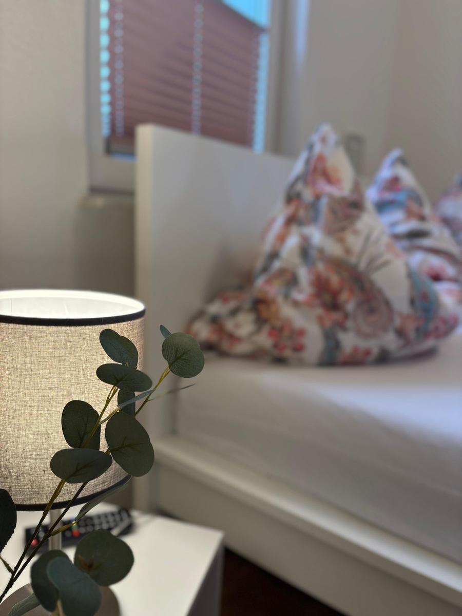 Schlafzimmer mit Bett, Nachttisch und Lampe. Fenster mit Jalousie im Hintergrund.