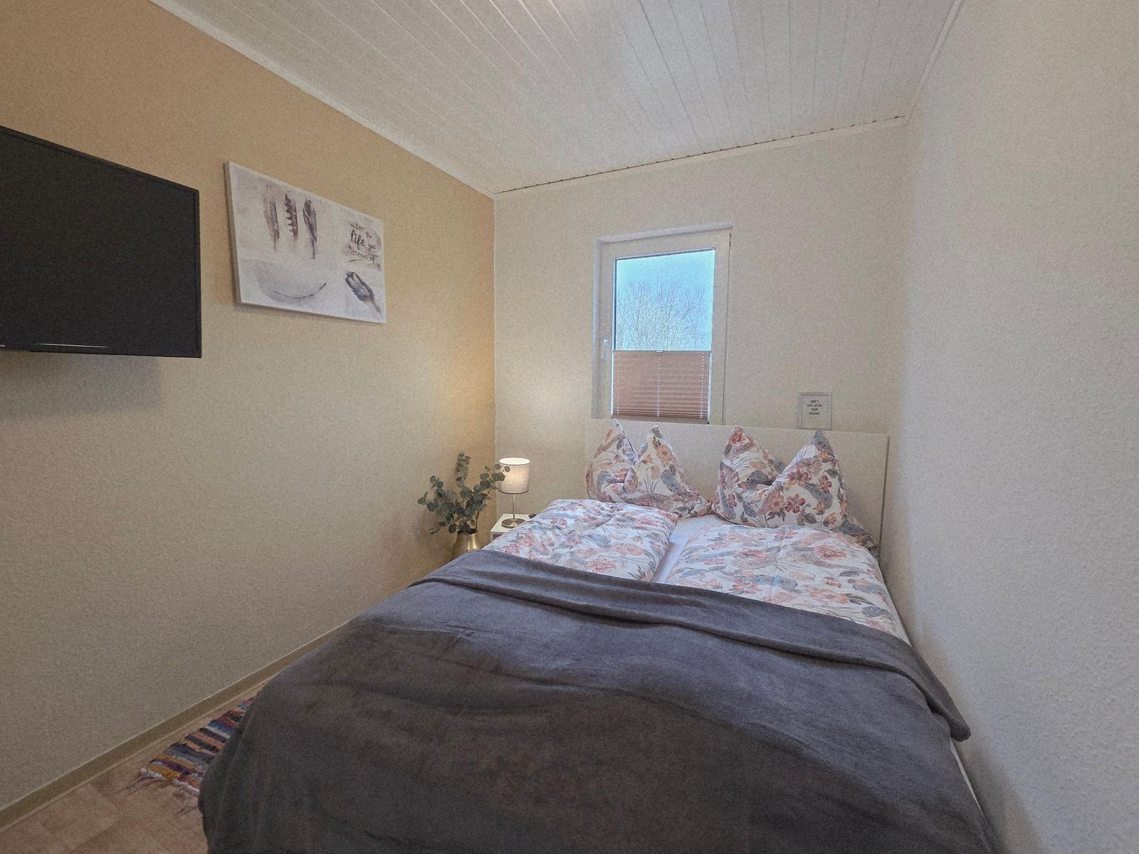 Schlafzimmer mit Bett, Fenster, Lampe und Wand-TV.