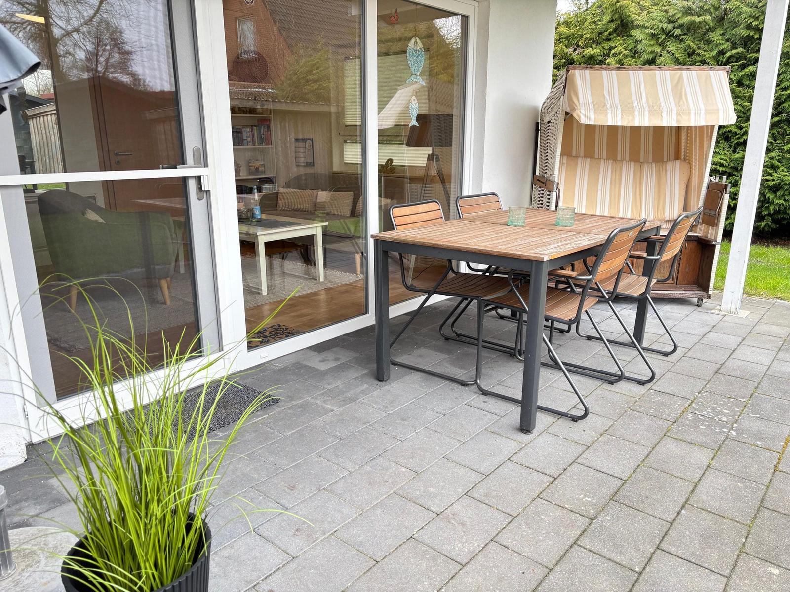 Terrasse mit Tisch, Stühlen und Strandkorb. Durchgang ins Haus mit Blick ins Wohnzimmer.