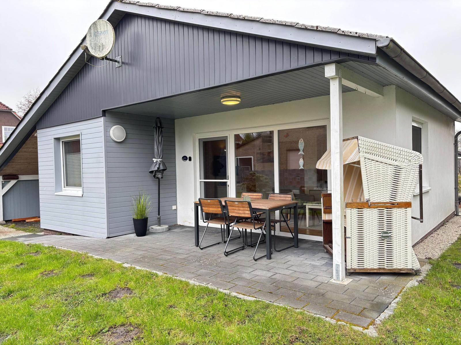 Haus mit Terrasse, Tisch, Stühlen und Strandkorb. Außenbereich mit Pflanze und Satellitenschüssel.