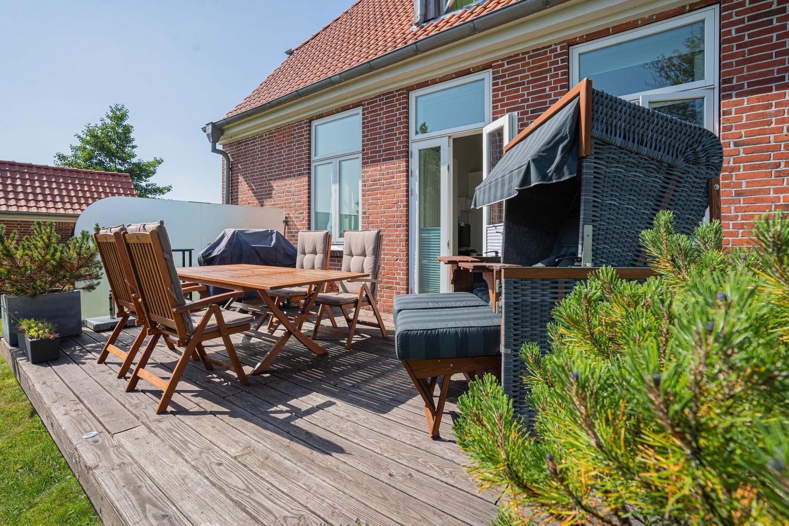 Terrasse mit Holztisch, Stühlen und Liege. Hintergrund: roter Ziegelbau mit Fenstern.