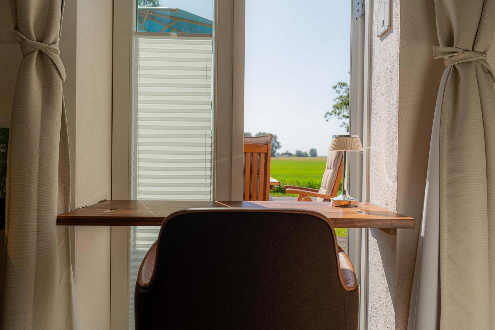 Schreibtisch mit Stuhl vor Fenster, Blick auf grünes Feld und Gartenmöbel