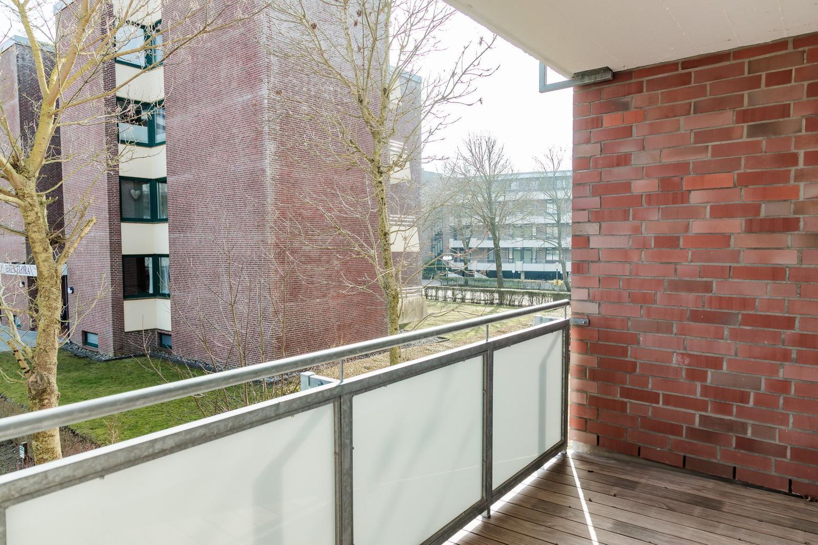 Terrasse mit Holzboden und Glasgeländer, Blick auf rote Backsteinhäuser und kahle Bäume.