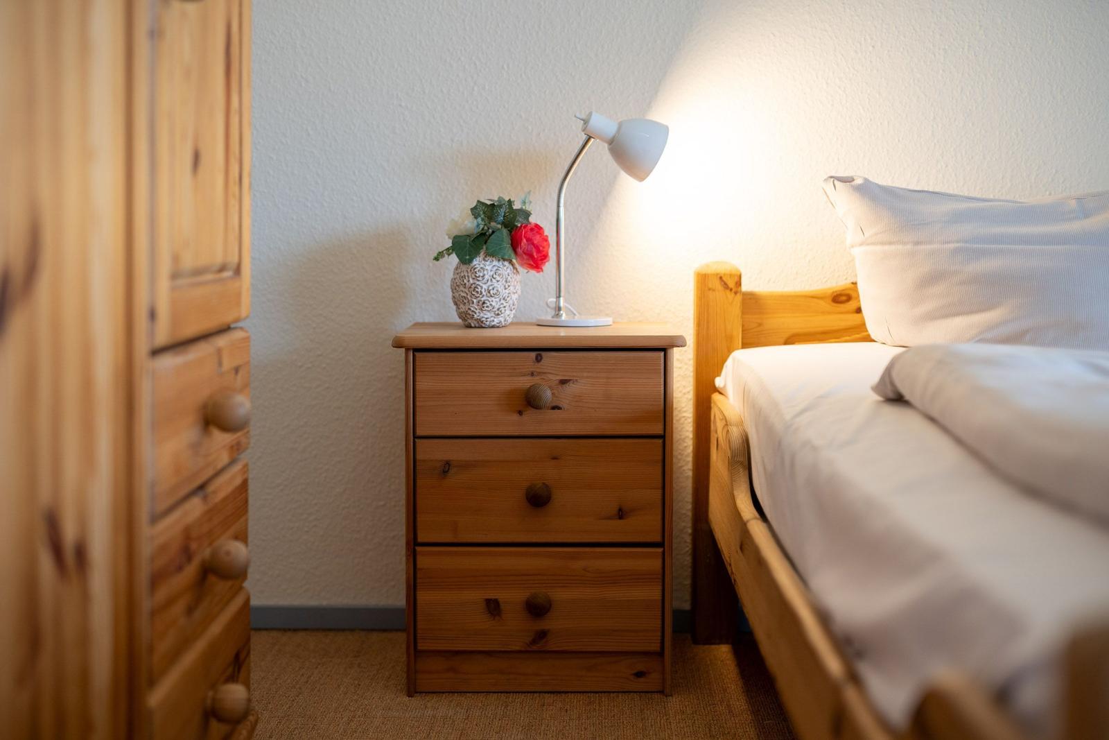 Schlafzimmer mit Holzbett, Nachttisch und Lampe. Auf dem Nachttisch steht eine Blumenvase.