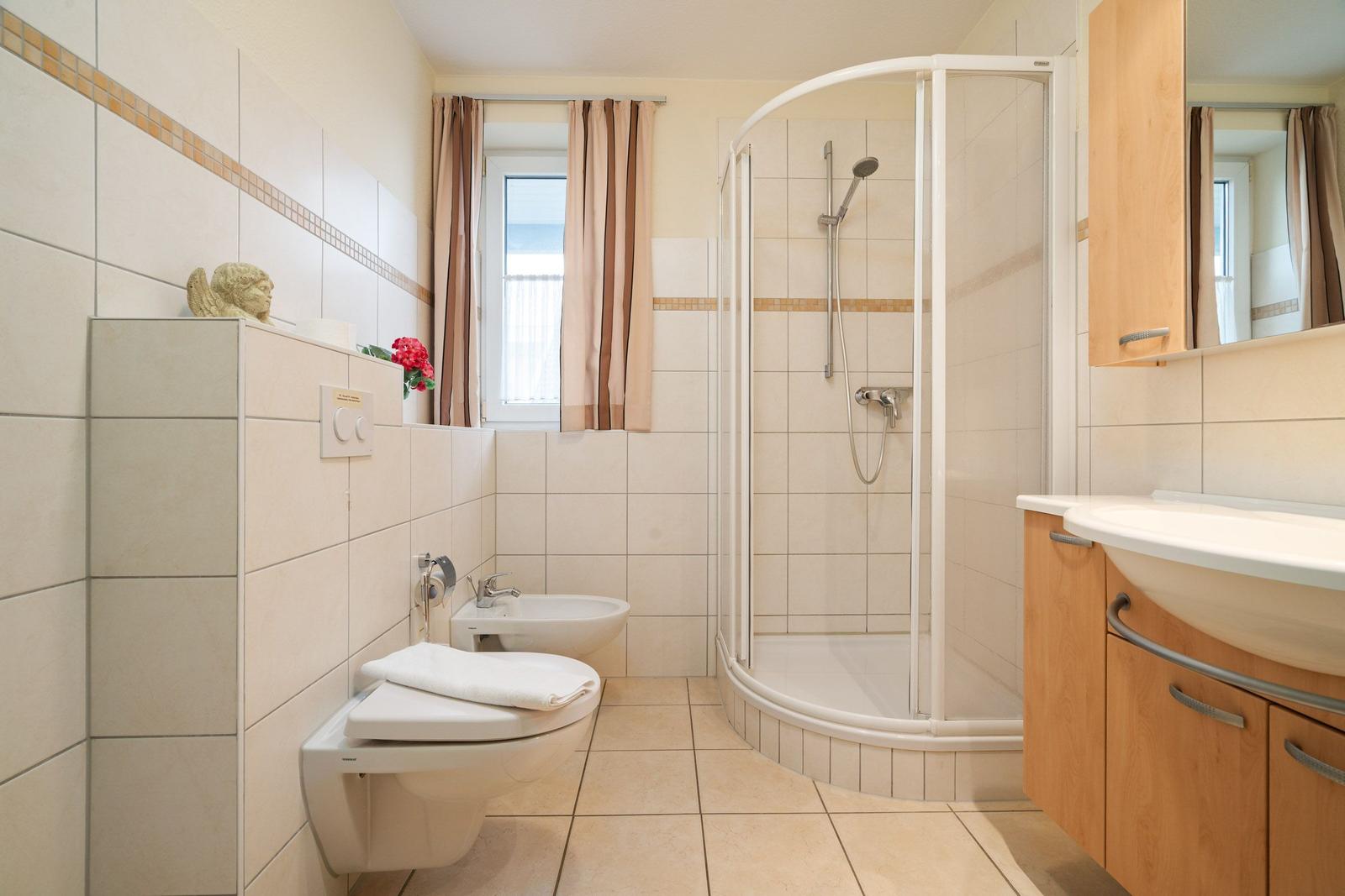 Badezimmer mit Dusche, Waschbecken und Toilette. Fenster mit Vorhängen.