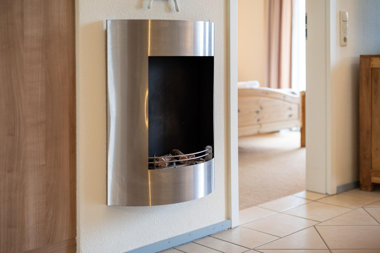 Wandmontierte Feuerstelle mit Holz im Flur. Im Hintergrund ist ein Schlafzimmer mit Bett sichtbar.
