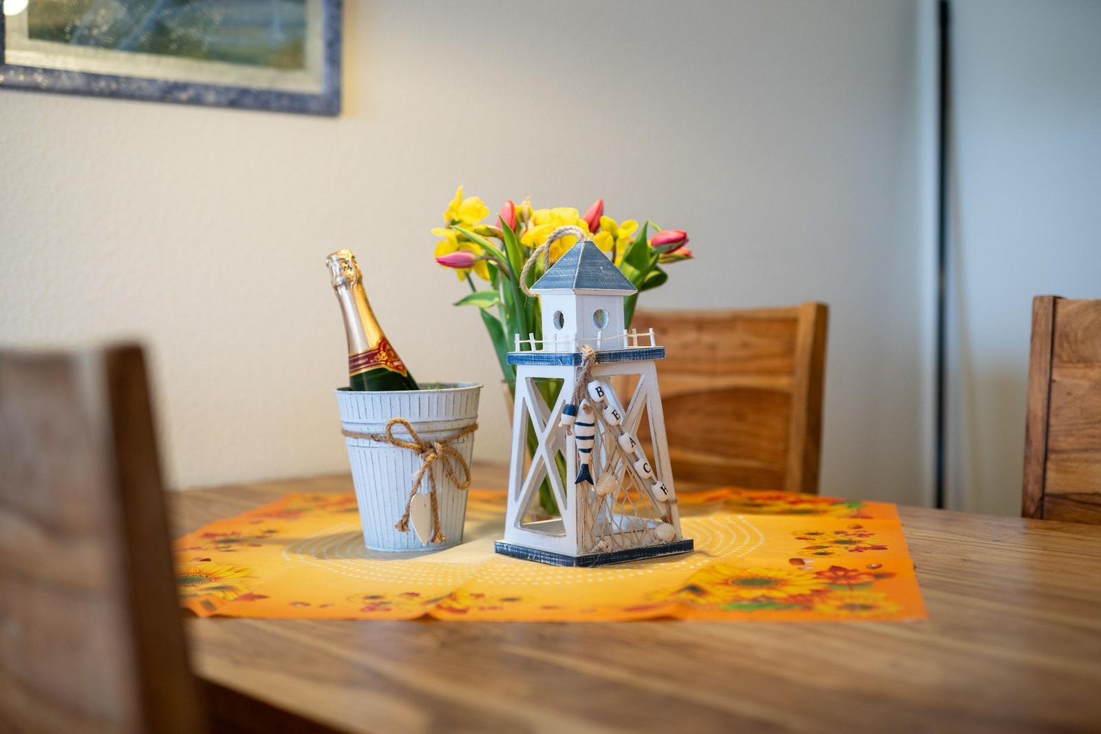 Holztisch mit Sonnenblumen-Tischdecke, Flasche Champagner und Strandturm-Dekoration.