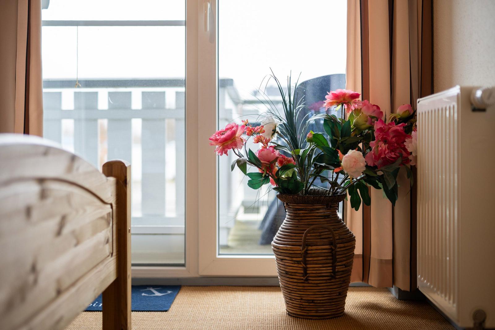 Ein Bett neben einem Fenster mit Blumen in einem Korb. Draußen ist eine Terrasse.