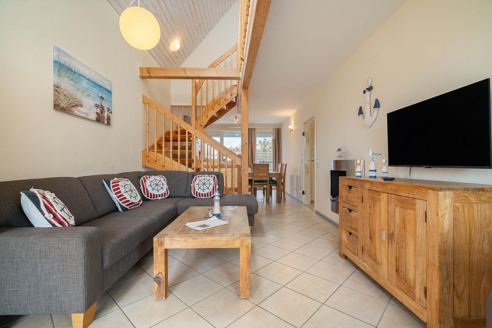 Wohnzimmer mit Sofa, Couchtisch und Holztreppenhaus. Fernseher und Küchenbereich sichtbar.