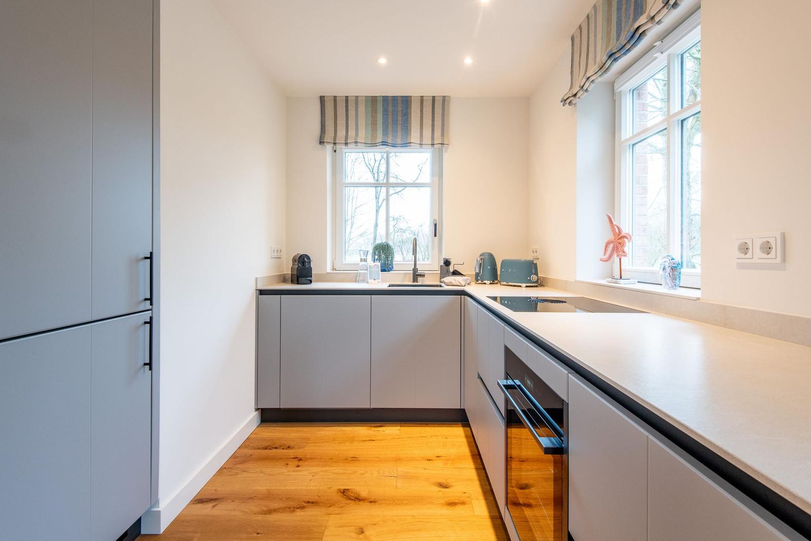 Küche mit grauen Schränken, Holzboden und Fenstern mit Rollläden.