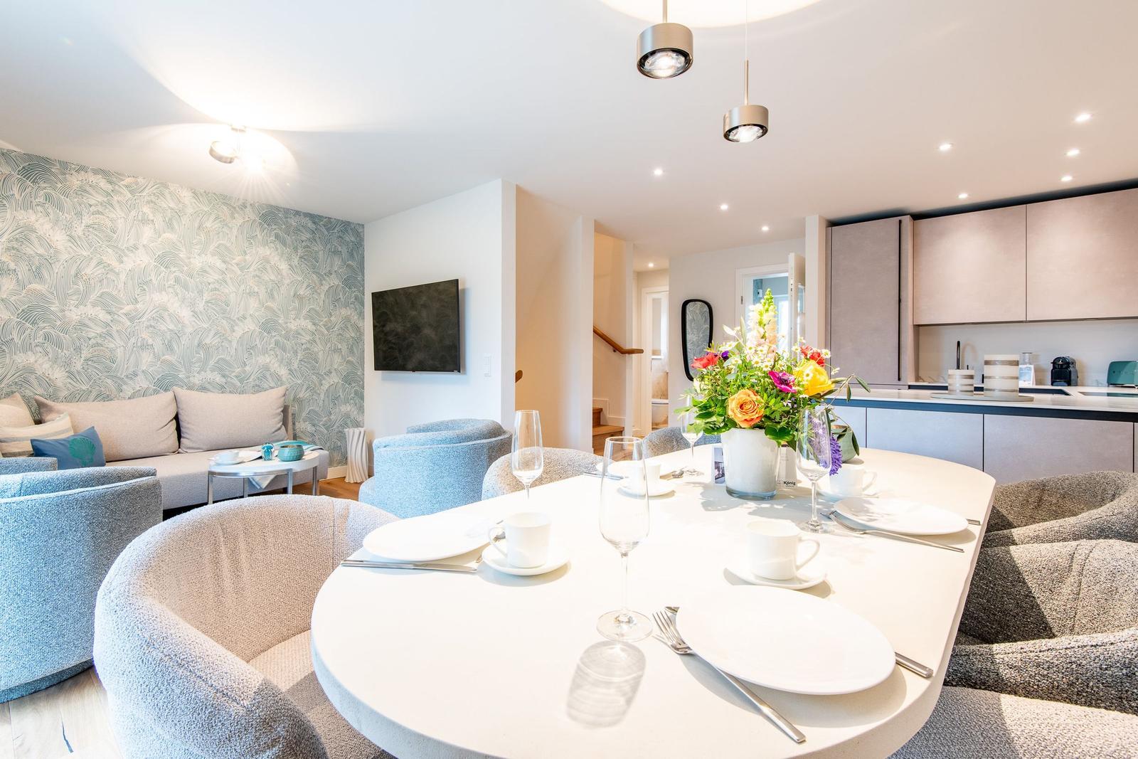 Esszimmer mit runder Tafel, Blumen und Sitzgelegenheiten. Küche und Wohnbereich im Hintergrund.