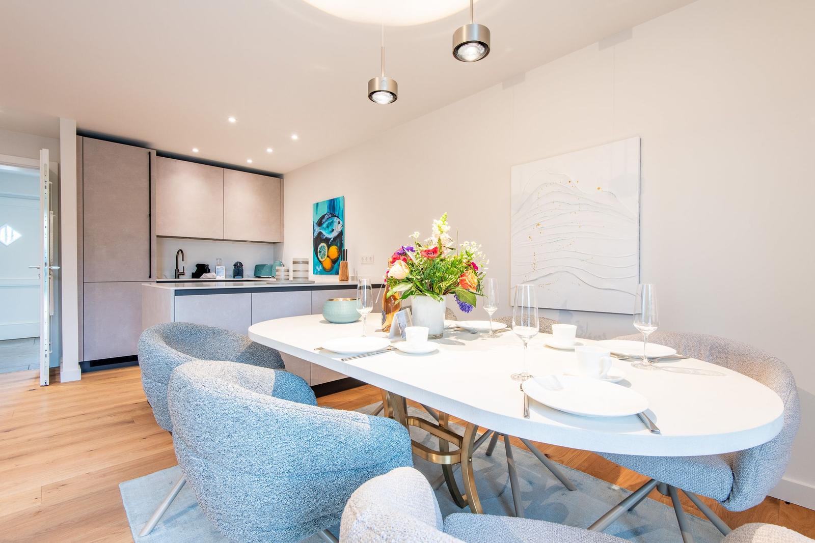 Essbereich mit Tisch und Stühlen, Küche im Hintergrund, Wandbilder und Blumen.