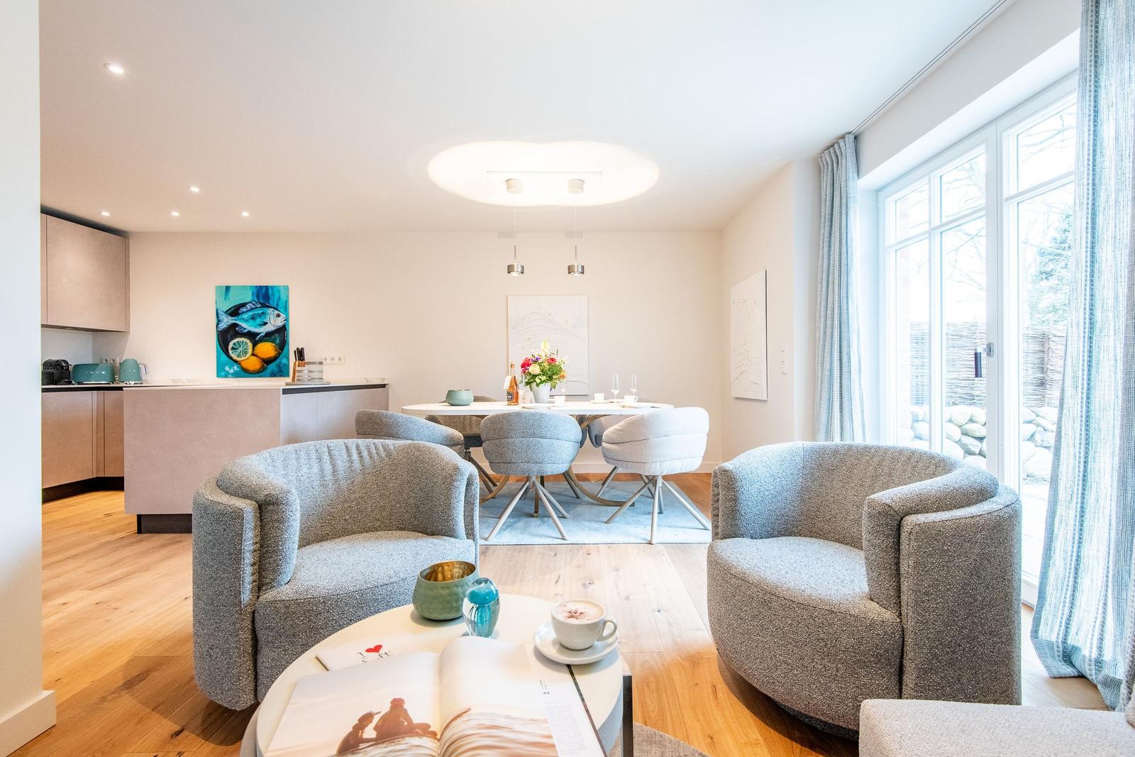 Wohn- und Essbereich mit Küchenzeile und Fenster. Zwei Sessel, Tisch, Blumen und Bild.