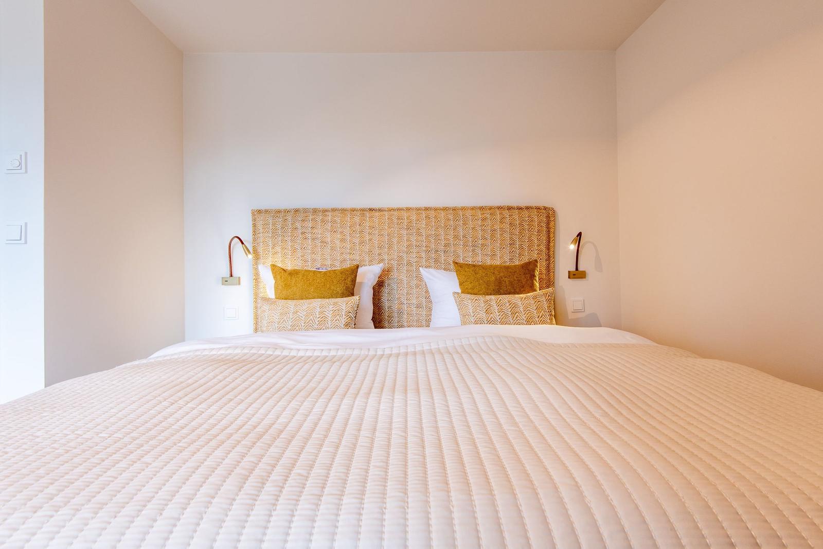 Ein Bett mit weißer Bettdecke und gelben Kissen. An den Wänden hängen Wandlampen.