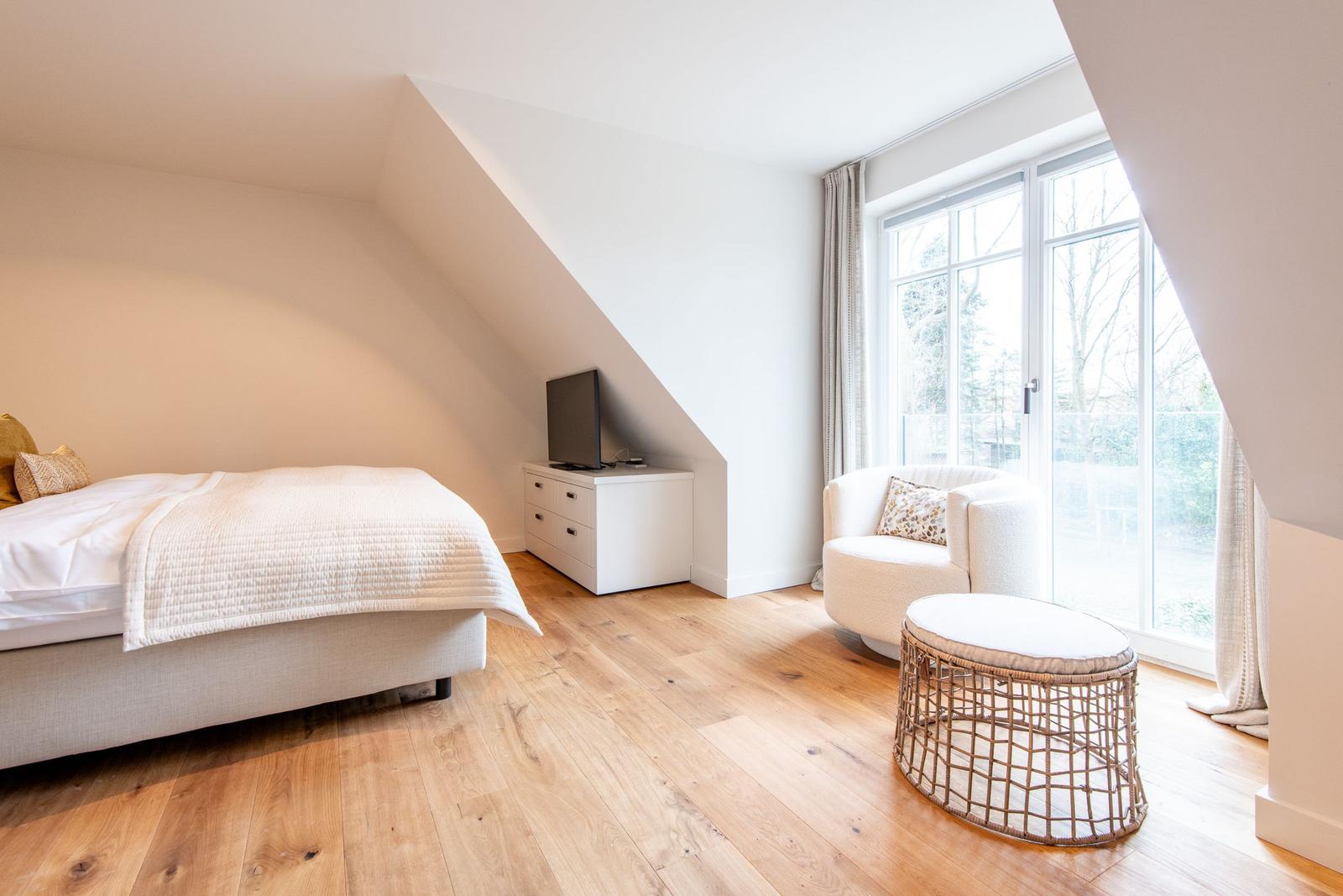 Schlafzimmer mit Bett, Fernseher und Sitzgelegenheit. Holzboden und großes Fenster.