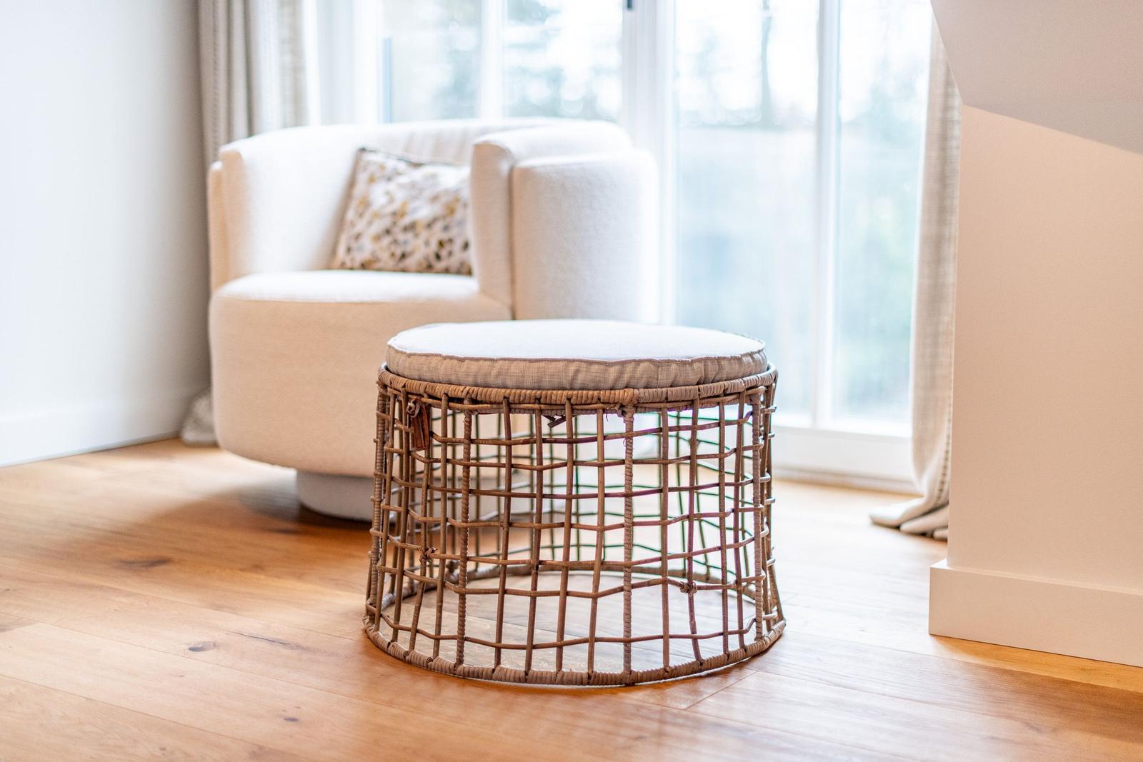Rattan-Sitzhocker mit Kissen vor einem Sessel und Fenster
