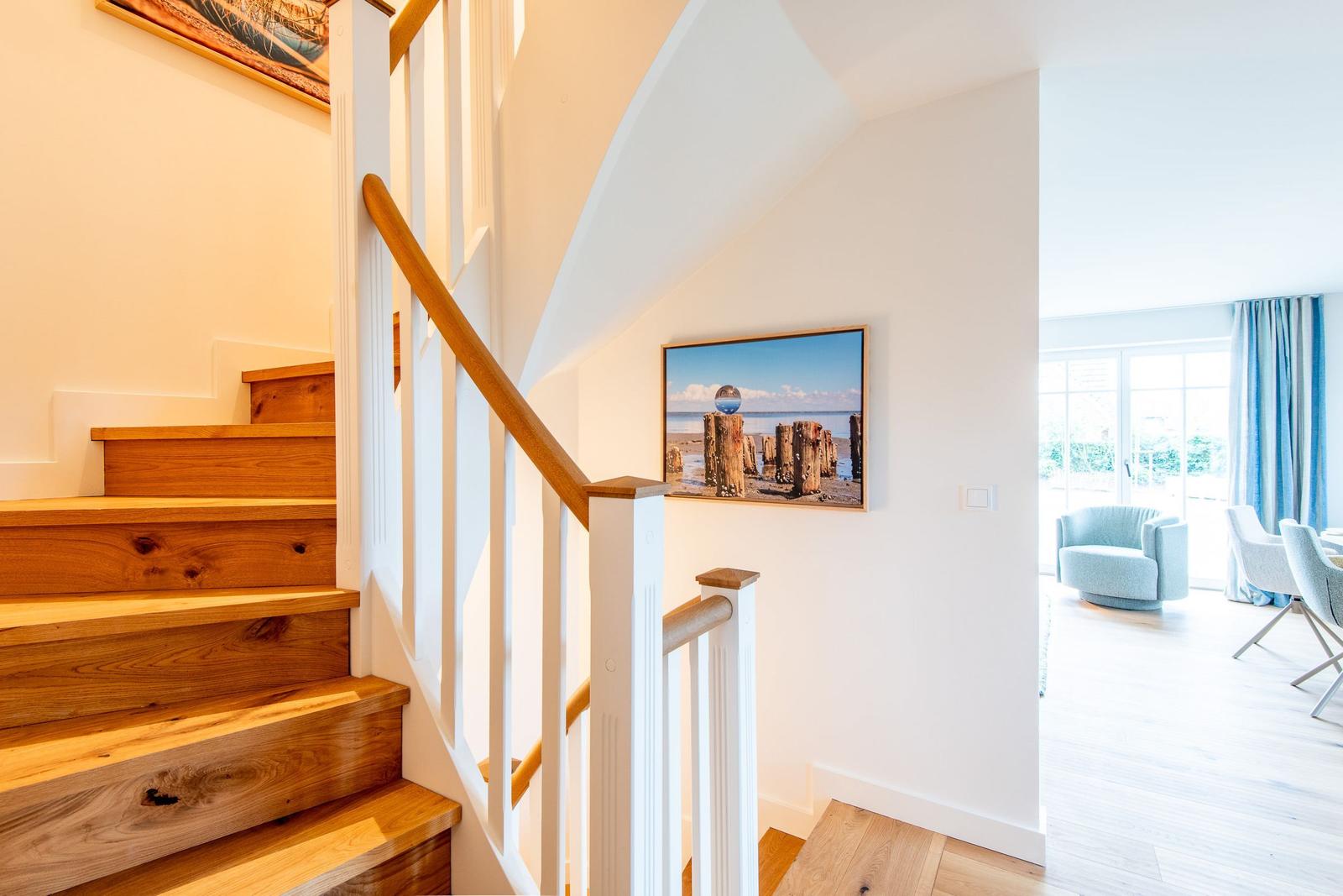 Holztreppenhaus mit Bild an der Wand und Blick in Wohnbereich mit Sitzgelegenheiten.