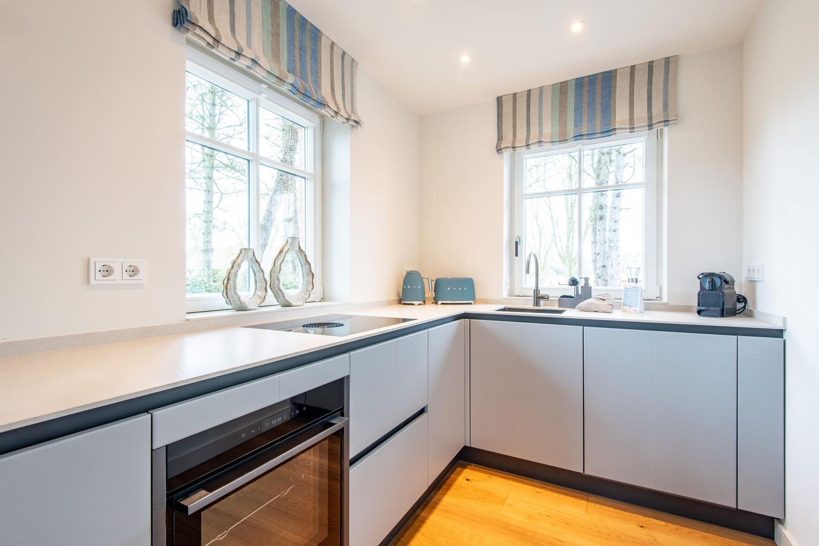 Küche mit hellen Schränken, Arbeitsplatte und zwei Fenstern mit Rollläden.