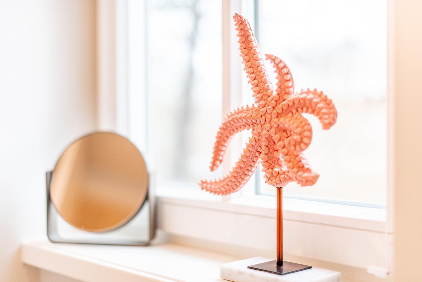 Ein orangefarbener Seestern auf einem Standfuß steht auf einem Fensterbrett neben einem runden Spiegel.
