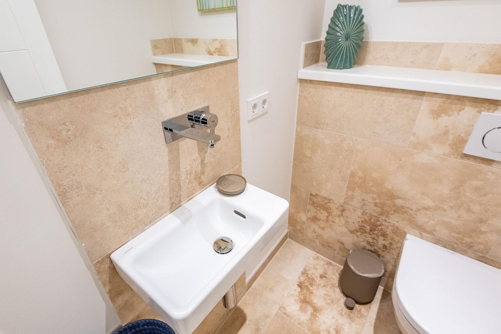 Badezimmer mit Waschbecken, Spiegel und Toilette. Wand und Boden sind mit hellen Fliesen ausgelegt.