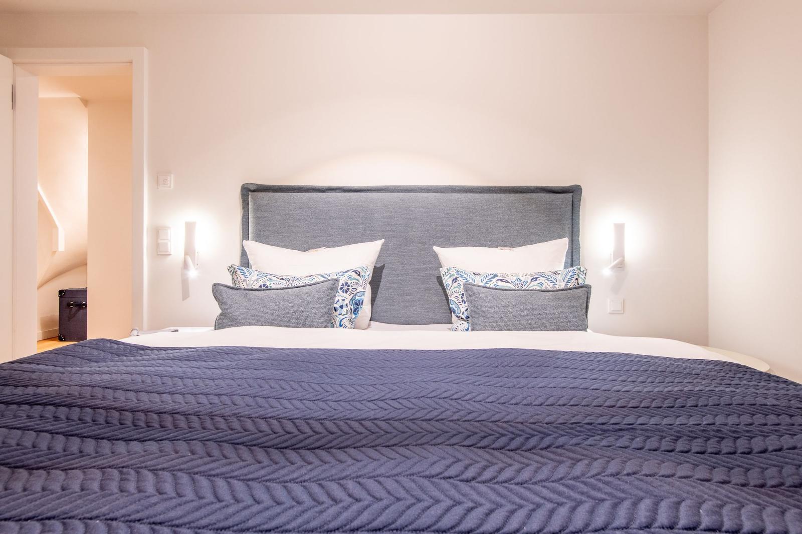 Doppelbett mit blauer Decke und Kissen, Wandlampen und Tür im Hintergrund.