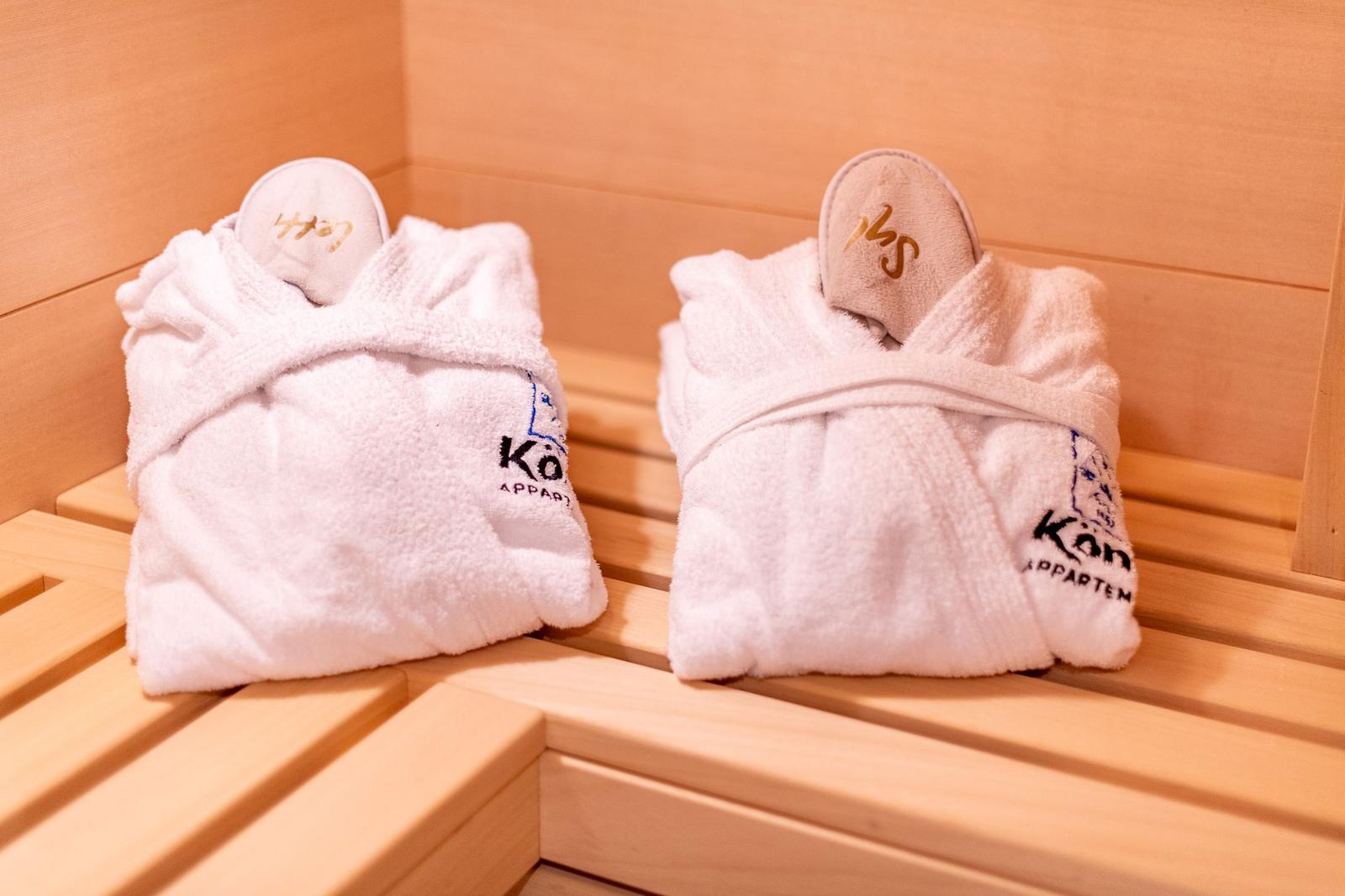Zwei weiße Bademäntel mit Schuhen liegen auf Holzbank in Sauna.