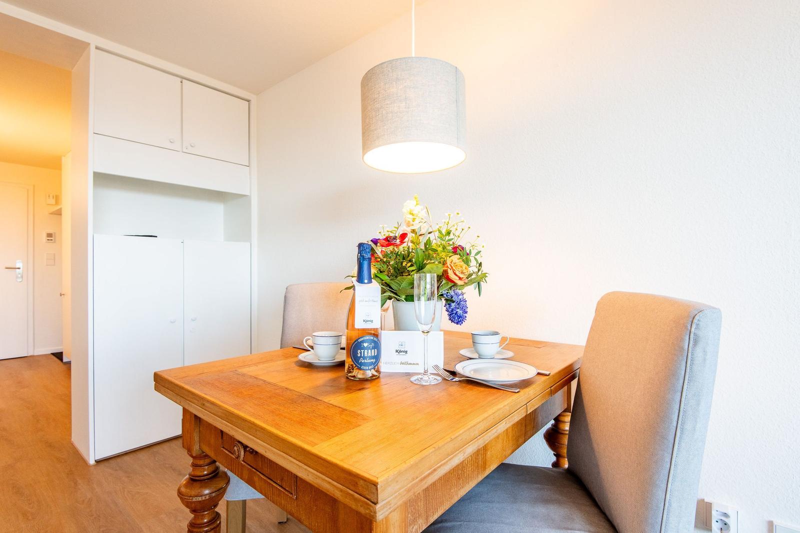 Esstisch mit Stühlen, Blumen und Flasche. Hintergrund: weiße Schränke und Deckenleuchte.